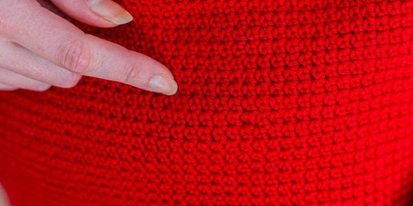 Une main à la peau pâle désigne un tissu au crochet d'un rouge vif, mettant en valeur la texture et le motif détaillés des mailles réalisées au point de rose au crochet.