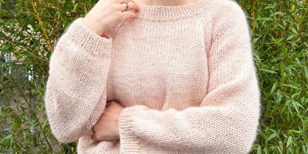 Une personne portant un pull tricoté rose pâle et doux - ressemblant à un modele tricot gratuit de pull oversize pour débutant - se tient devant des plantes de bambou vertes, souriant et regardant sur le côté, une main touchant son cou.