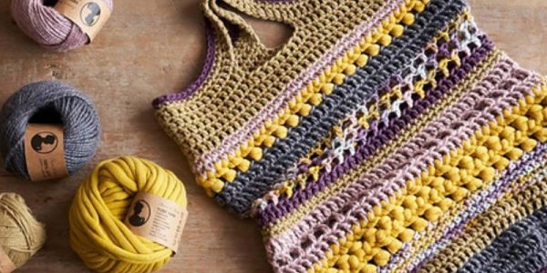Un tote bag au crochet aux couleurs vives, avec des rayures jaunes, grises, roses, blanches et violettes, repose sur une surface en bois à côté de quatre pelotes de laine : une source d'inspiration idéale pour votre prochain projet de sac fourre-tout au crochet.