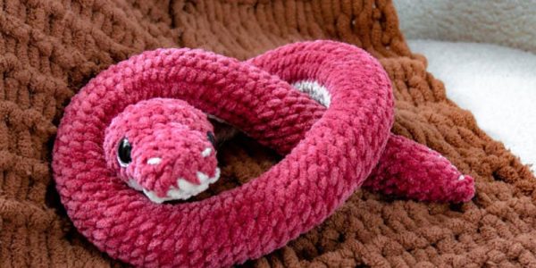 Un serpent en peluche rose, tricoté, avec des détails blancs, repose sur une couverture marron texturée, mettant en valeur le charme des tutoriels d'amigurumis au crochet.