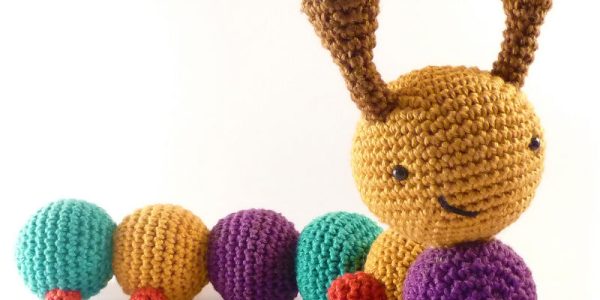 Une chenille au crochet avec une tête jaune, des antennes brunes, des segments de corps bleus et violets, et de petites pattes rouges sourit sur un fond blanc - parfait pour suivre un tuto chenille au crochet gratuit.