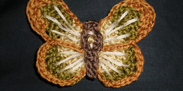 Un papillon au crochet, réalisé avec du fil marron, vert, beige et orange, repose sur un fond de tissu foncé. Ses ailes présentent des couleurs superposées et un motif texturé, ce qui en fait un exemple charmant pour ceux qui recherchent des tutoriels d’appliqués d’animaux au crochet.