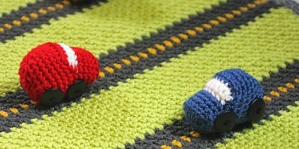 Trois petites voitures au crochet — rouge, jaune et bleue — sont disposées sur un tapis de jeu crocheté vert, gris et jaune.